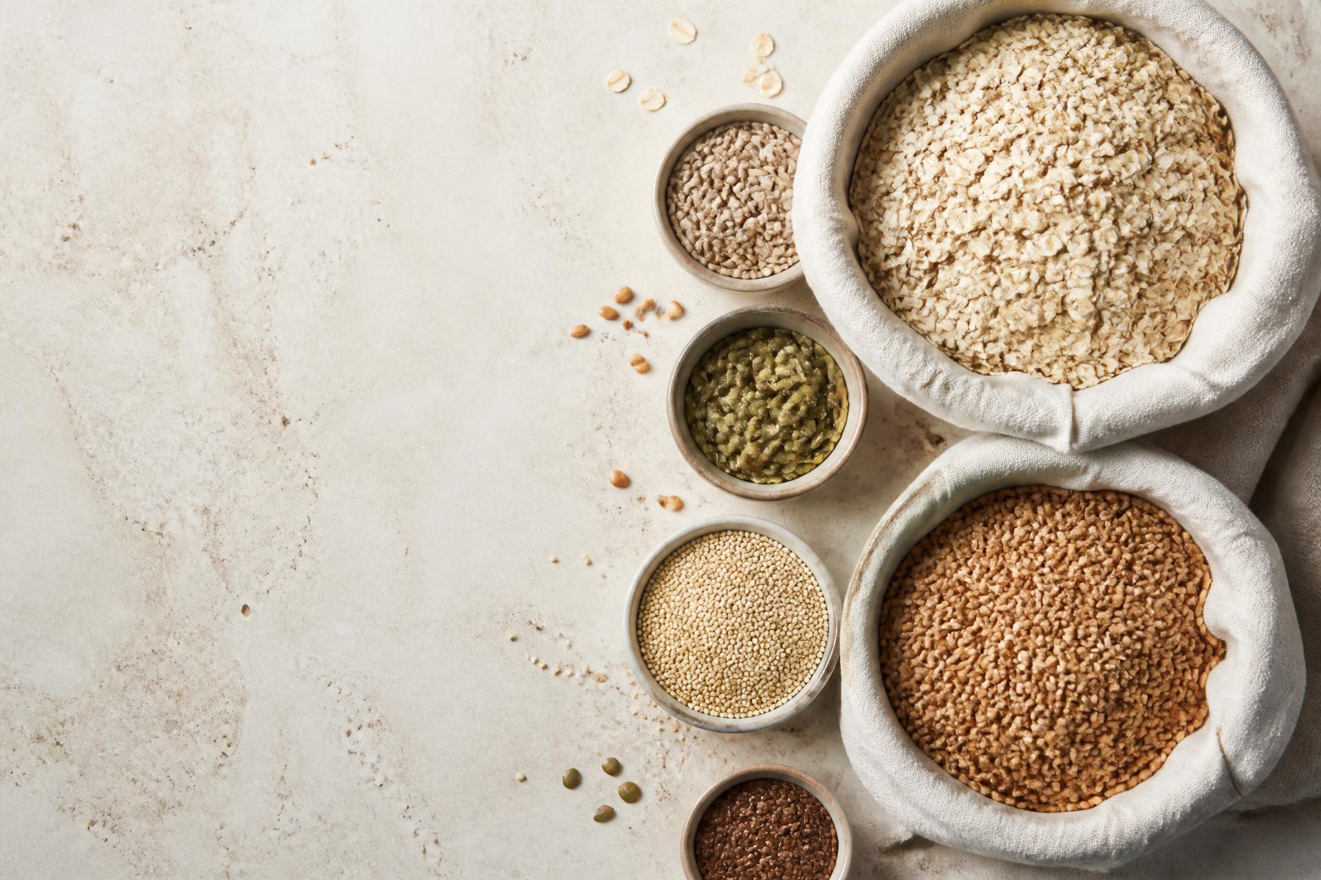 Overhead view of oats, grains, and seeds arranged in linen sacks and ceramic bowls on a light stone surface
